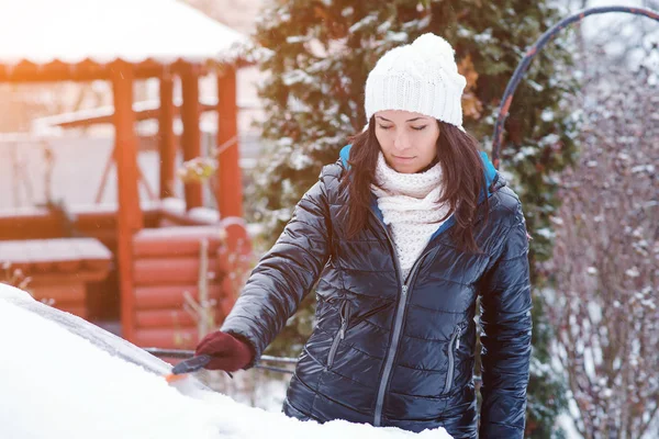 Young attractive woman in winter clothes removes snow from car with snow brush. — Stock Photo, Image