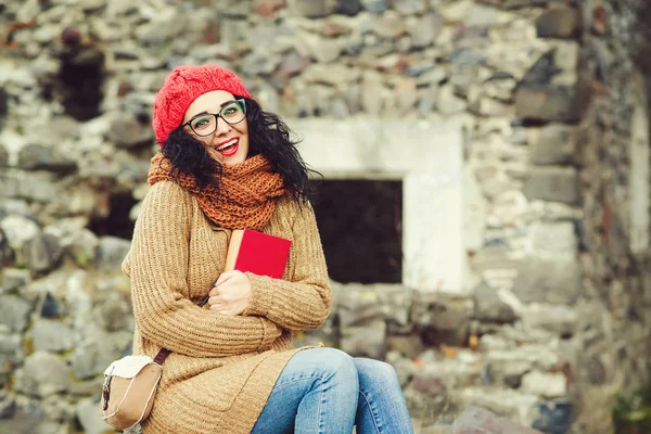 Cheerful young woman in red knitted hat with a book, looking to camera and laughing outdoor. — Stock Photo, Image