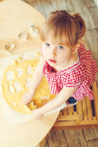 Cute little daughter is cutting cookie dough in different shapes. Daughter helps her mother bake cookies in a kitchen. — Stock Photo, Image