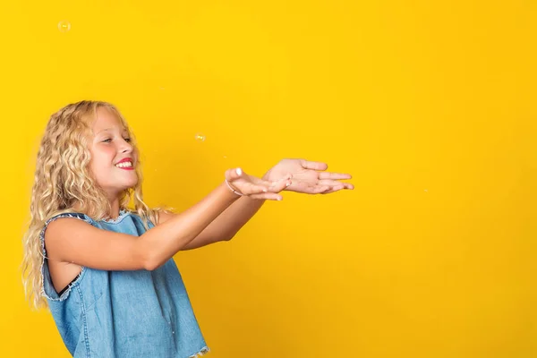 Adorável menina loira brincando com bolhas de sabão sobre um fundo amarelo . — Fotografia de Stock