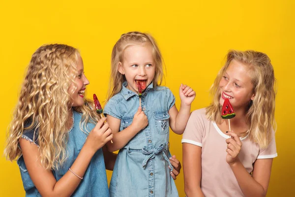 Šťastné děti jíst ovoce lízátka v studio, roztomilé dívky sestry či přátelé baví společně izolované žluté pozadí. — Stock fotografie