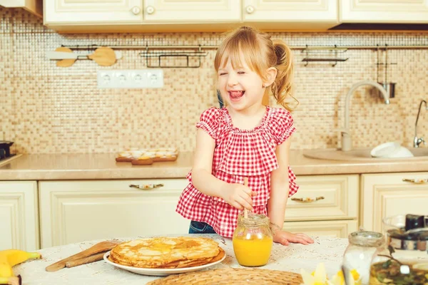 Schattig klein meisje bereiden pannenkoeken en gieten honing op stapel pannenkoeken. — Stockfoto