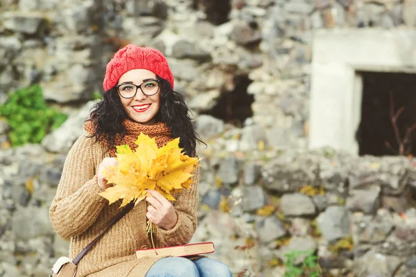 Adorable young woman in red knitted hat holding bouquet of maple leaves and enjoying beautiful autumn. — Stock Photo, Image