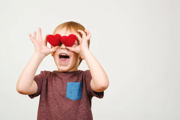 Leuke speelse jongen zijn ogen achter gebreide rode harten, geïsoleerd op een witte muur, copyspace verbergen. — Stockfoto