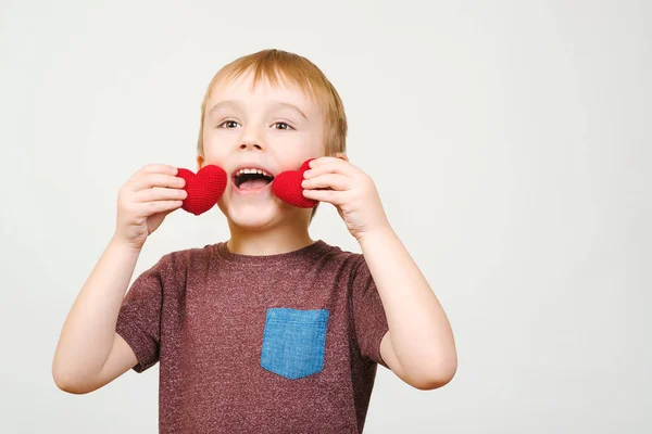Aranyos boldog fiú játszik két kötött piros szívek, elszigetelt fehér falon, copyspace. — Stock Fotó