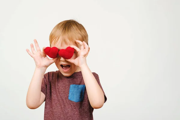 Leuke speelse jongen zijn ogen achter gebreide rode harten, geïsoleerd op een witte muur, copyspace verbergen. — Stockfoto