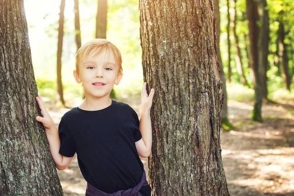 Feliz chico lindo apoyado contra el árbol y sonriendo durante caminar en un bosque . — Foto de Stock