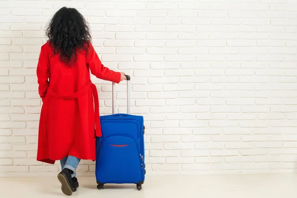 Вид сзади на стильную женщину в красном пальто с чемоданом на стене из белого кирпича . — стоковое фото