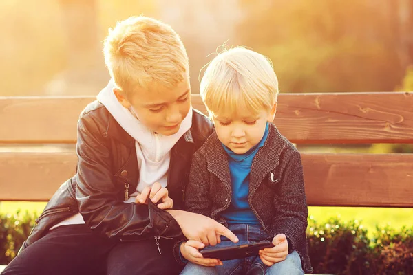 Az idősebb testvér megtanítja a fiatalabbat a mobiltelefon használatára. Gyerekek ülnek a padon.. — Stock Fotó