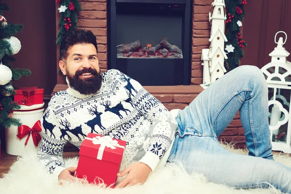 Glad skäggig man vilar nära öppen spis vid jul. Mannen i stickad tröja sitter framför skorstenen. — Stockfoto