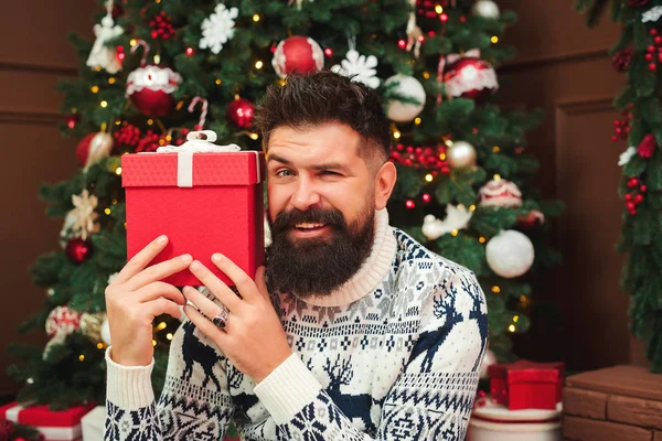 Handsome bearded guy holding christmas gift. Excited happy man in christmas sweater sitting near christmas tree.