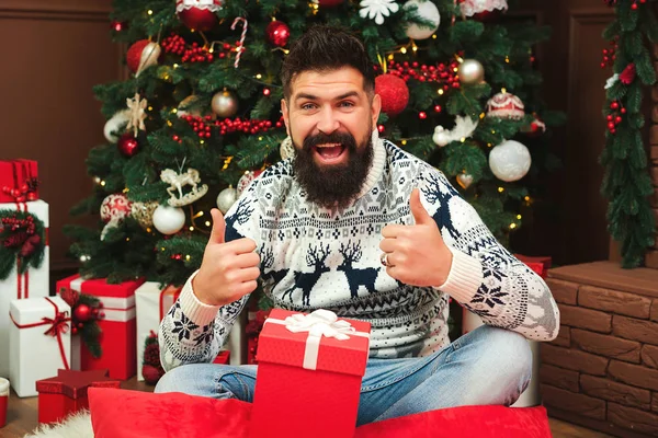 Yılbaşı kazaklı sakallı adam Noel ağacının yanında oturuyor. Moda adamı el kol hareketi yapar. — Stok fotoğraf