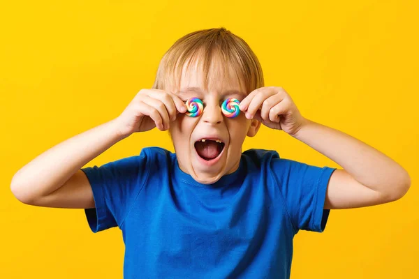 Criança feliz cobrindo os olhos com dois pirulitos coloridos. Bonito menino animado sobre fundo amarelo — Fotografia de Stock