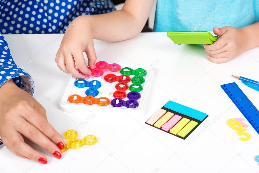 Mother with kid plays logical games, close up, top view. Child training mind with plastic colored puzzles on white table