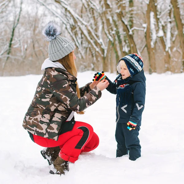 Anya segít a fiának kesztyűt húzni télen. A család téli sétán van a havas parkban. Téli ruhák gyerekeknek — Stock Fotó