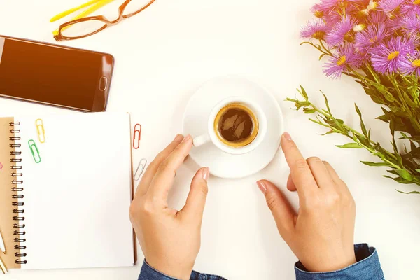 Жінка тримає чашку кави на білому офісному столі. Чистий блокнот, окуляри та весняна квітка на столі — стокове фото