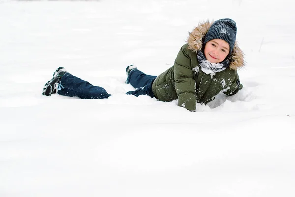 Boldog fiú fekszik a hóban a téli parkban. Téli alsónemű gyerekeknek. Érzelmes gyerek játszik a hóban a szabadban. Boldog téli ünnepeket! — Stock Fotó