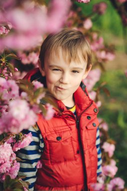 Stylish boy making grimace over spring nature background. Sakura tree blooming. Handsome kid on a walk in spring park