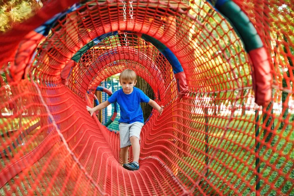 Cute child boy overcomes obstacles in rope tunnel outdoors. Modern amusement park for kids. Healthy and happy childhood. — Stock Photo, Image