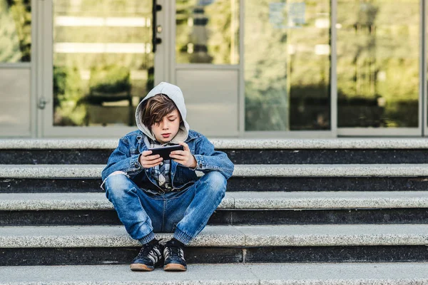 Мальчик играет в мобильные игры по телефону. Подросток со смартфоном сидит на лестнице на улице. Концепция интернет-зависимости. Подросток носит джинсовую куртку — стоковое фото