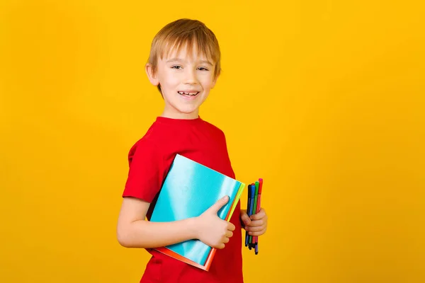 Ragazzo felice che tiene rifornimenti scolastici su sfondo giallo. Ragazzo con quaderni e penne. Concetto di ritorno a scuola — Foto Stock