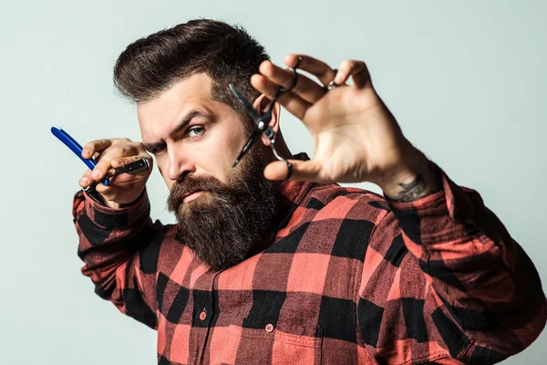 Peluquero con tijeras y navaja de afeitar. Peluquería pequeña empresa. Tipo barbudo con peinado elegante sobre fondo azul — Foto de Stock