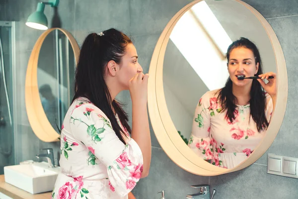 Hermosa mujer cepillándose los dientes por la mañana. Chica mirándose en el espejo en el baño. Interior moderno de baño. Cuidado dental y concepto de salud —  Fotos de Stock