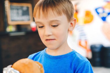 Hungry cute boy holds a burger at outdoors cafe. Happy child eating fast food. Childhood, unhealthy food concept.