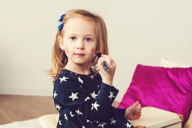Küçük kız annesinin kozmetiğiyle oynuyor. Evde makyaj yapan genç bir kız. Kız makyajla eğleniyor. Mutlu bir çocukluk. Çocuk kozmetik ürünleri. Çocuk modası, yaşam tarzı ve eğlence..