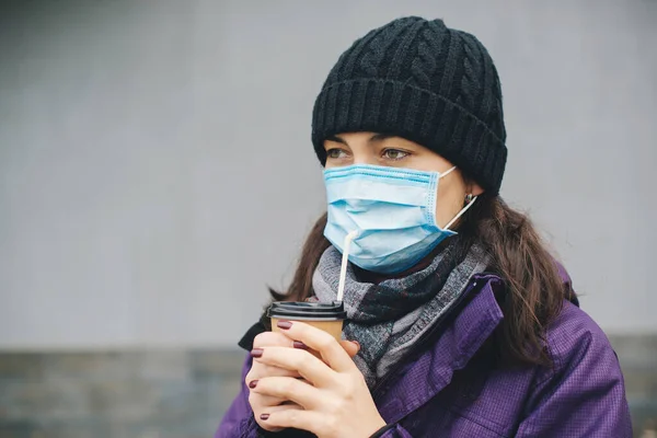 Face mask protective for spreading of coronavirus. COVID-19 pandemic coronavirus. Woman in city street wearing face mask. Woman with surgical mask drinks coffee outdoors. Coronavirus epidemic in city.