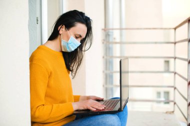 Kadın yüz maskesi. Balkonda dizüstü bilgisayarda çalışan bir kız. Teknoloji, eğlence ve yaşam tarzı. Freelancer, uzaktan kumanda. Coronavirus karantinası. Evden çalışıyorum. Karantina sırasında uzaktan eğitim.