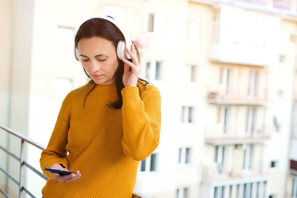 Young woman with headphones breathing fresh air on a balcony. Woman using mobile phone and wireless headphones. Girl listen music at balcony. Stay home. Self isolation at home. Coronavirus quarantine.