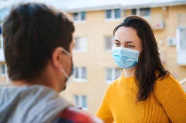 Genç çift dışarıda balkonda konuşuyor. Koronavirüs karantinası sırasında evde izolasyon. Yüz maskesi takan kadın ve erkek. Coronavirus koruma ve önleme. Evde kal. Balkonda dikilen çift.