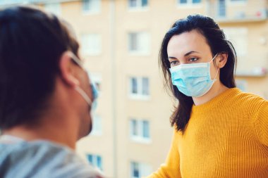 Genç çift dışarıda balkonda konuşuyor. Koronavirüs karantinası sırasında evde izolasyon. Yüz maskesi takan kadın ve erkek. Coronavirus koruma ve önleme. Evde kal. Balkonda dikilen çift.