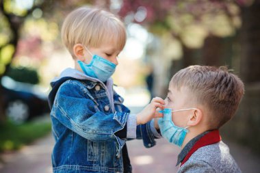 Dışarıda tıbbi maske takan çocuklar. Oğlan, küçük erkek kardeşine yüzüne maske takmasına yardım eder. Bahar zamanı yürüyüşe çıkan bir aile. Coronavirüs salgını. Koronavirüsü önleme. Güvenlik maskeli çocuklar.