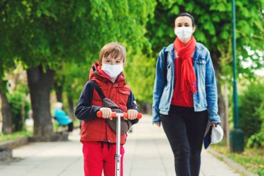 Dışarıda maskeli bir aile var. Parkta scooter süren bir çocuk. Çocuk tıbbi maske takıyor. Gerçek hayat 2020. Coronavirüs salgını. Anne ve oğlu koornavirüs karantinası sırasında yürüyüşe çıktılar..