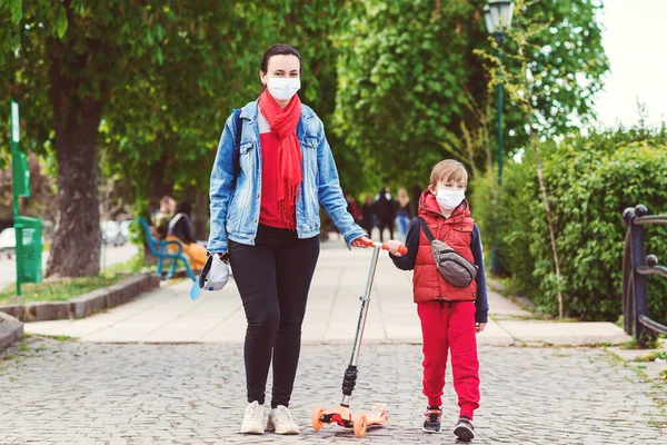 Child in face mask riding on scooter in street. Boy wears medical mask. Family in safety masks outdoors. Real life 2020. Coronavirus epidemic. Mother and son on a walk during coornavirus quarantine.