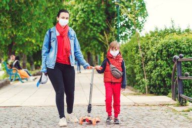 Dışarıda maskeli bir aile var. Parkta scooter süren bir çocuk. Çocuk tıbbi maske takıyor. Gerçek hayat 2020. Coronavirüs salgını. Anne ve oğlu koornavirüs karantinası sırasında yürüyüşe çıktılar..
