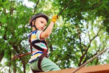 Çocuk dağ miğferi ve güvenlik ekipmanlarıyla parka gidiyor. Yüksek tel parka tırmanma macerası. Çocuklar için yaz kampı. Küçük çocuk ağaçların arasından geçen kablo yolundan geçiyor..