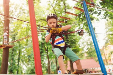 Çocuk dağ miğferi ve güvenlik ekipmanlarıyla parka gidiyor. Yüksek tel parka tırmanma macerası. Çocuklar için yaz kampı. Küçük çocuk ağaçların arasından geçen kablo yolundan geçiyor..