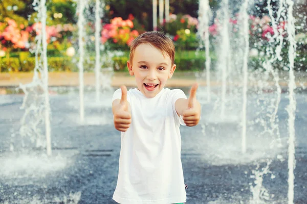 Boy is cooled by water from the fountain. Hot sunny summer day. Happy kid playing at a fountain with water. Stylish boy near fountain. Child showing thumbs up outdoors. Summer vacation, lifestyle.