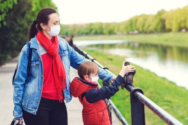 Dışarıda maskeli bir aile var. Parkta scooter süren bir çocuk. Çocuk tıbbi maske takıyor. Gerçek hayat 2020. Coronavirüs salgını. Anne ve oğlu koornavirüs karantinası sırasında yürüyüşe çıktılar..