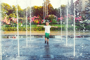 Şehir parkında su akışı arasında koşan mutlu çocuk. Çocuk çeşmeden gelen suyla serinliyor. Mutlu çocuk, Synny yaz gününde çeşmedeki su jetlerinin arasında oynuyor. Yaz tatili, yaşam tarzı..