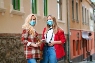 Coronavirus tecridinden sonra yaz tatili. Turist kızlar sokakta maske takıyor. Yeni şehri keşfeden arkadaşlar. Seyahat 2020. Yaşam tarzı, arkadaşlık ve tatiller. Eski şehirde yürüyen kızlar.