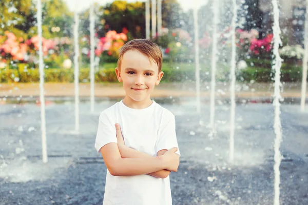 Portrait of happy boy over fountain background. Summer vacation, lifestyle. Boy is cooled by water from the fountain. Hot sunny summer day. Happy kid playing at a fountain with water.