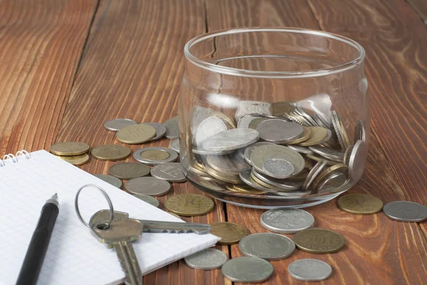 Strona główna oszczędności, koncepcja budżetu. Notatnik, długopis, klucze i monety w szklanym słoju na stół drewniany office. — Zdjęcie stockowe