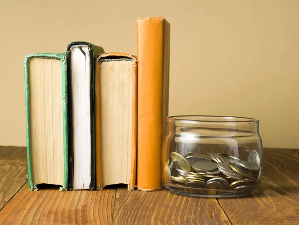 Κέρματα σε γυάλινο βάζο και στοίβα των βιβλίων πάνω ξύλινο τραπέζι. Εξοικονόμηση, έννοια financiai και εκπαίδευση. — Φωτογραφία Αρχείου