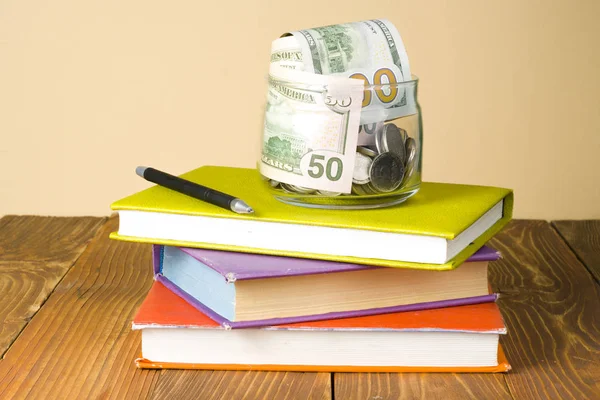 Χρήματα σε γυάλινο βάζο στη στοίβα των βιβλίων πάνω ξύλινο τραπέζι. Εξοικονόμηση, οικονομικών και Παιδείας έννοια. — Φωτογραφία Αρχείου