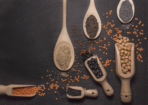 Sesame, chia and poppy seeds, red and black lentils, chickpeas and black beans with wooden spoons on black background
