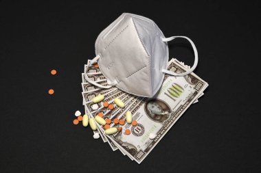 COVID-19 salgın konseptinin maliyeti ffp2 veya KN95 önleme maskesi, ilaçlar ve siyah arkaplan üzerine 100 dolarlık banknotlar.